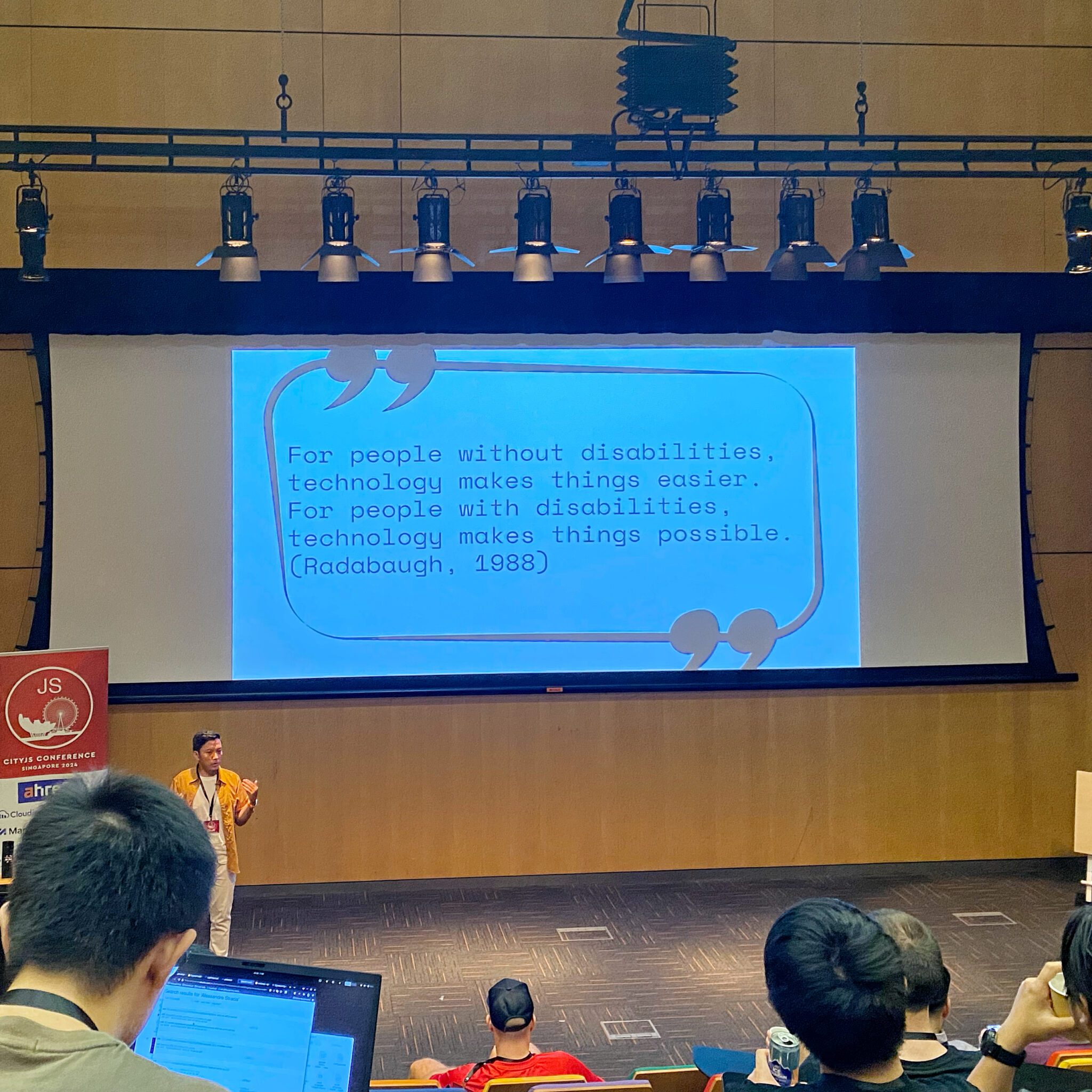 A developer from Indonesia is giving a talk about web accessibility. The text on the slider in the photo says: For people without disabilities, technology make things easier. For people with disabilities, technology make things possible. (Quote from Rabadabaugh, 1988)